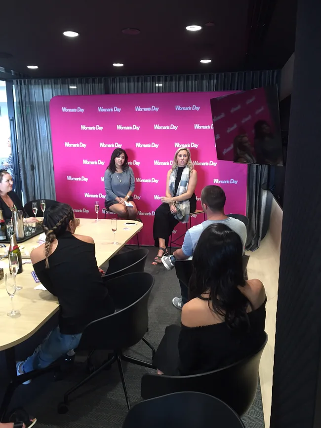 Women seated in discussion at a Women's Day event with a pink backdrop, surrounded by an audience.
