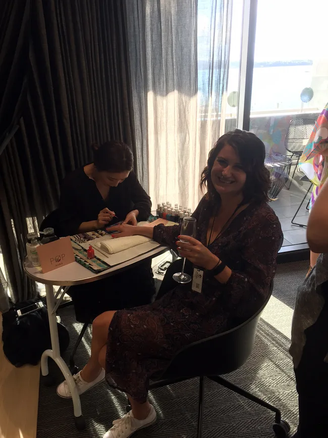 A woman sits smiling with a drink while getting a manicure near a window with light coming through sheer curtains.