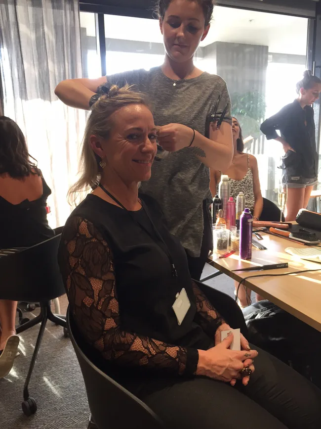 A woman smiles as a hairstylist works on her hair; salon tools are visible on a table.
