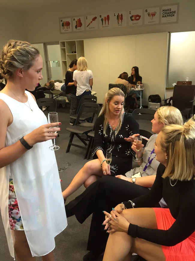 Group of women socializing and holding drinks in a casual setting with a motivational sign on the wall.