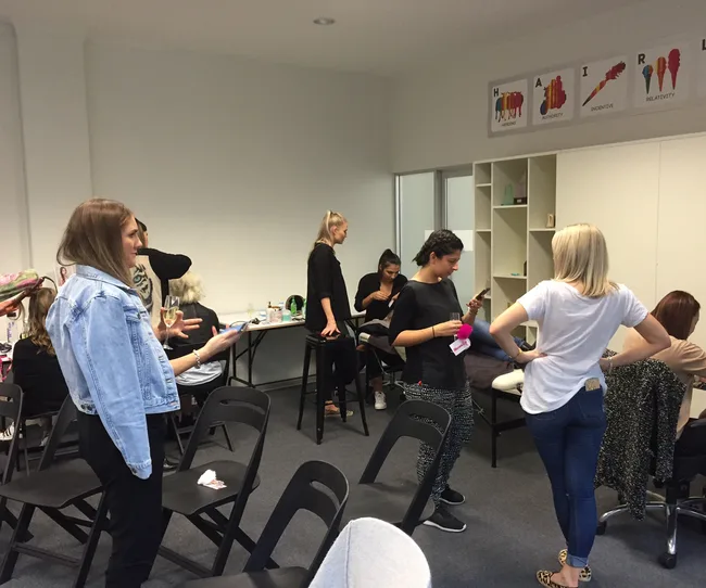 Group of people in a room interacting, some standing, others sitting, with hair styling posters on the wall.