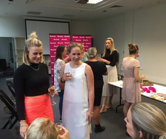 Women chatting and holding drinks at a social event in a room with a "Woman's Day" backdrop.