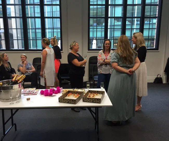 A group of people socializing in a room with windows, with a table of snacks and champagne in the foreground.