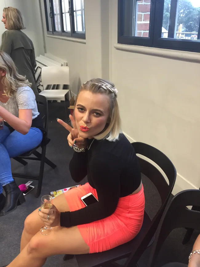 Young woman with braided blonde hair showing a peace sign while seated, wearing a black top and pink skirt, holding a drink.
