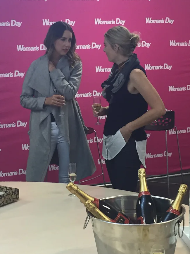 Two women talking at an event with a "Woman's Day" backdrop and champagne bottles in an ice bucket in the foreground.