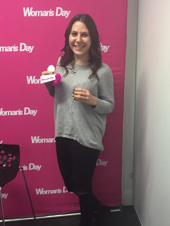 Woman smiling, holding a champagne flute and a pink-handled item, standing in front of a pink "Woman's Day" backdrop.