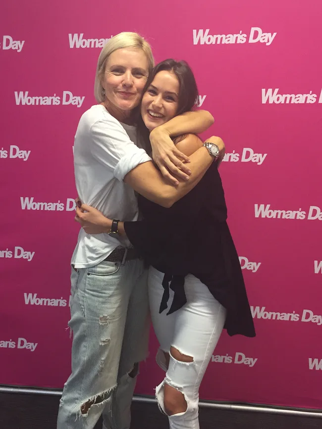 Two people smiling and hugging in front of a pink "Woman's Day" backdrop.