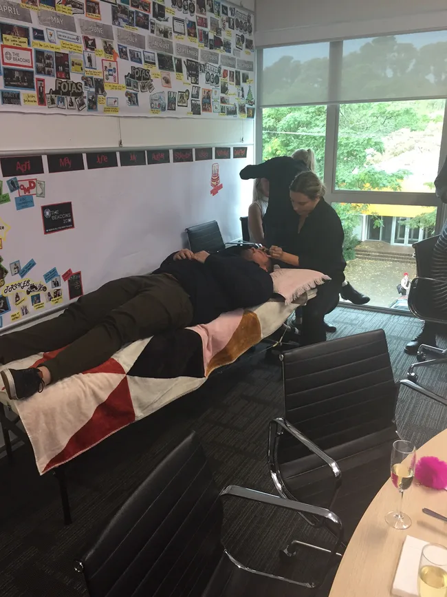 Person lying on a makeshift bed in an office while another person performs an eyebrow treatment.