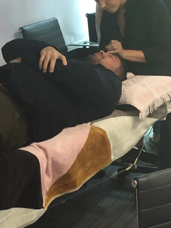 Man lying on a table getting eyebrows groomed by a person in a dark shirt.