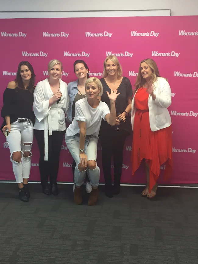 Group of six women posing in front of a pink "Woman's Day" backdrop, some smiling and waving.