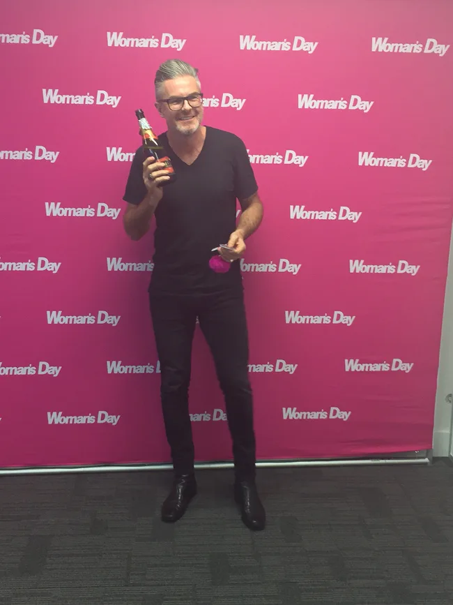 Man smiling, holding a drink, in front of a pink "Woman's Day" backdrop.