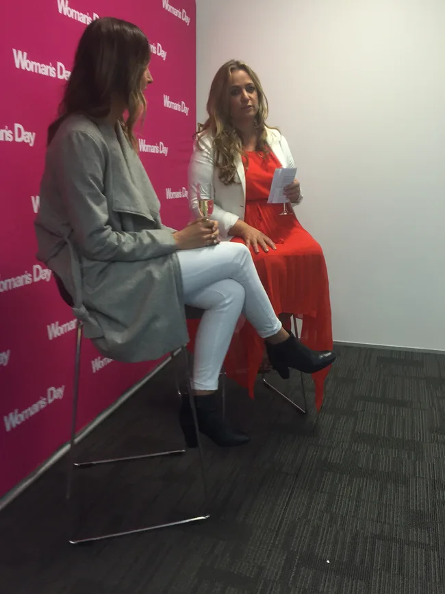 Two women seated on chairs at a Woman's Day event, one holding a drink, the other papers.