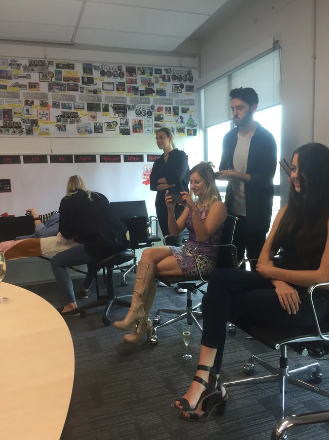 Group of people in an office meeting room, some seated and others standing, with a wall collage in the background.