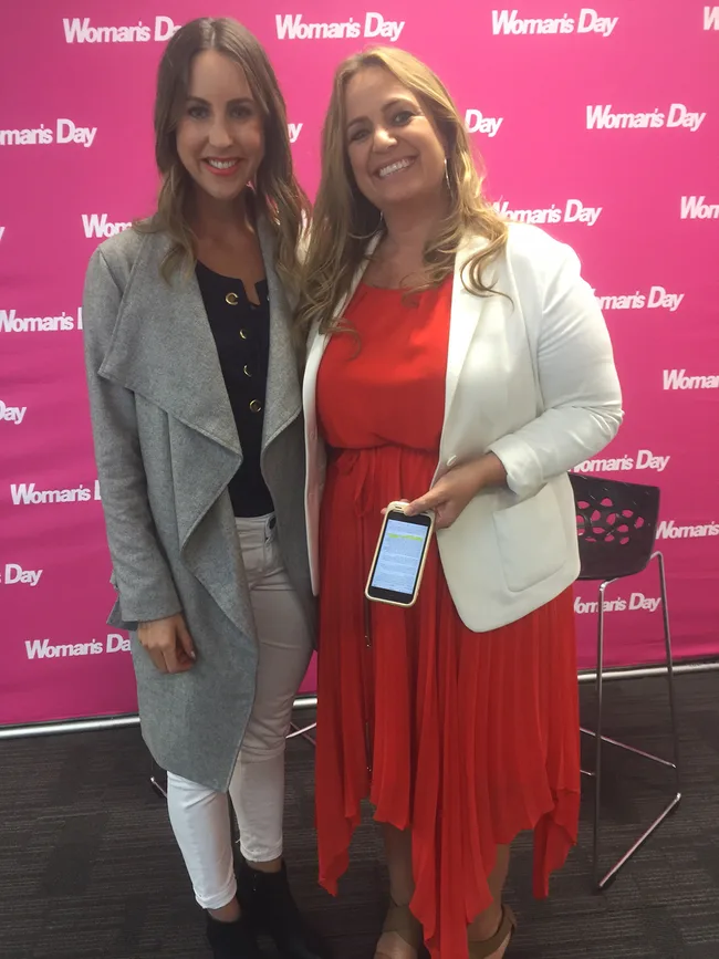 Two women posing in front of a pink "Woman's Day" backdrop; one holds a phone with text visible.