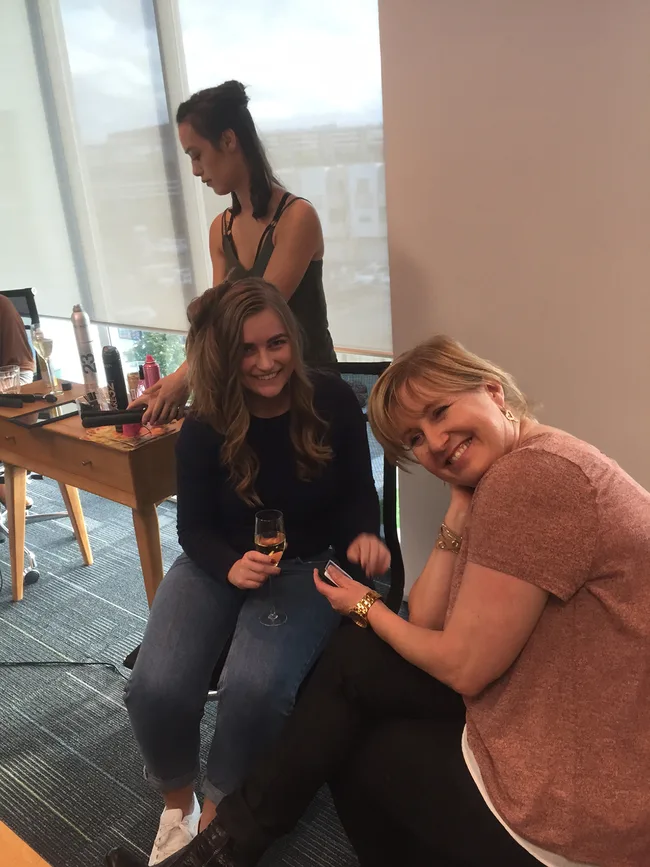 Three women in a room, one styling hair with curling iron, another sitting and smiling, holding champagne.