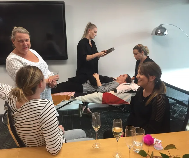 A group of women enjoying a spa treatment session in an office setting, with one woman receiving a facial.