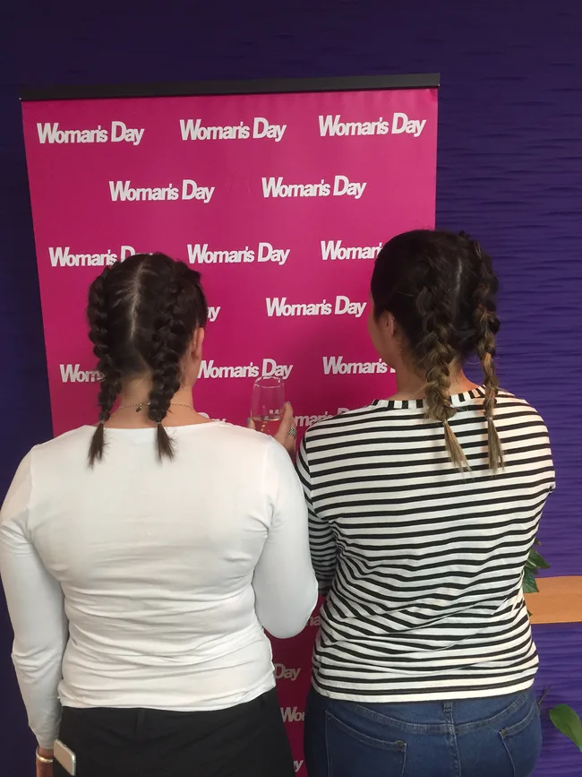 Two women with braided hair facing a "Woman's Day" backdrop, one holding a glass.