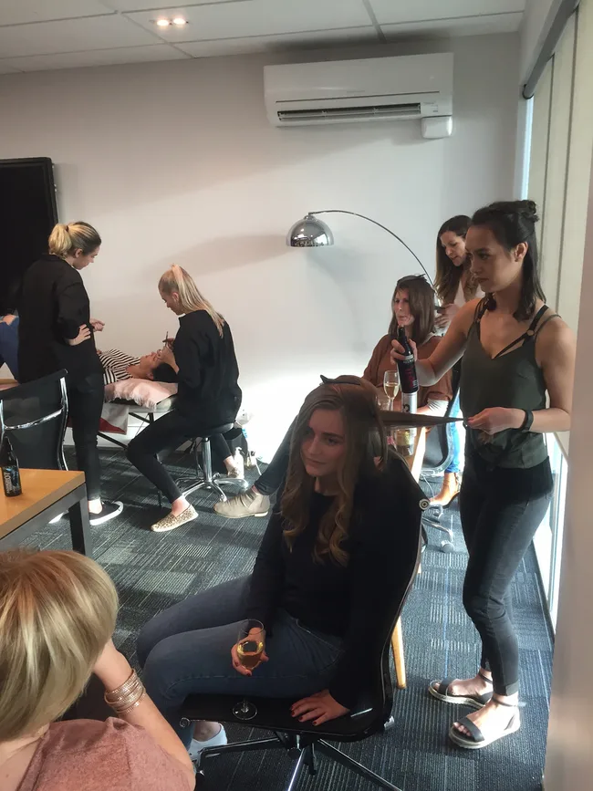 Group in a room, hairstylist working on a woman's hair while others chat and relax, one holding a drink.