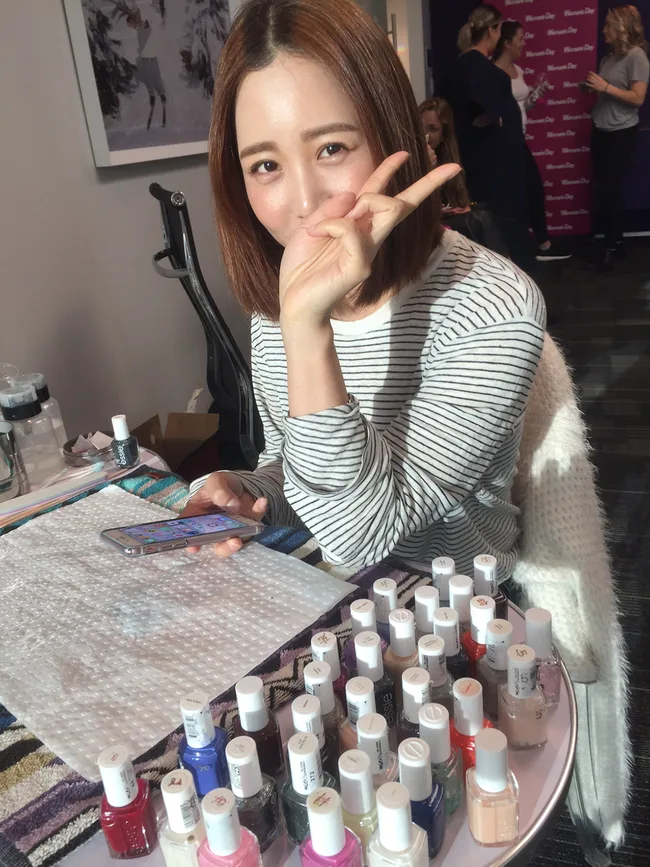 Young woman at a table with nail polishes, making a peace sign and holding a phone.