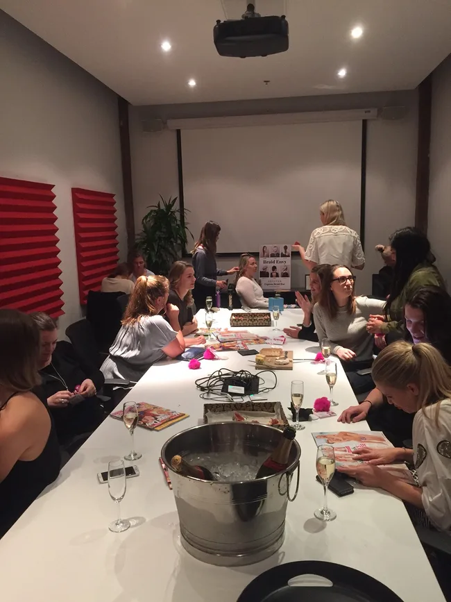 Group of people in a meeting room with magazines and drinks on a long table; one holds a "Braid Envy" booklet.