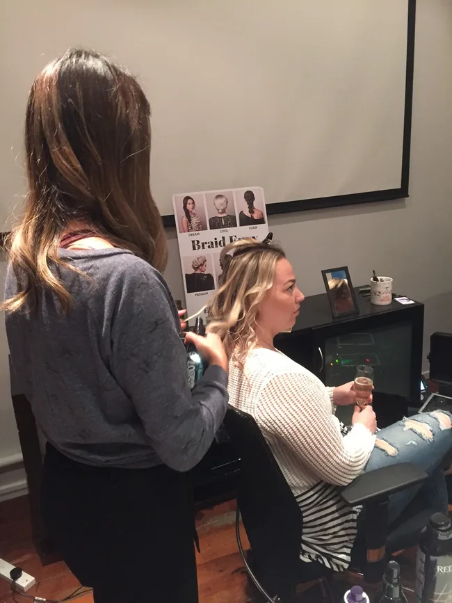 A woman styles another woman's hair in a salon, with a braid style chart visible in the background.