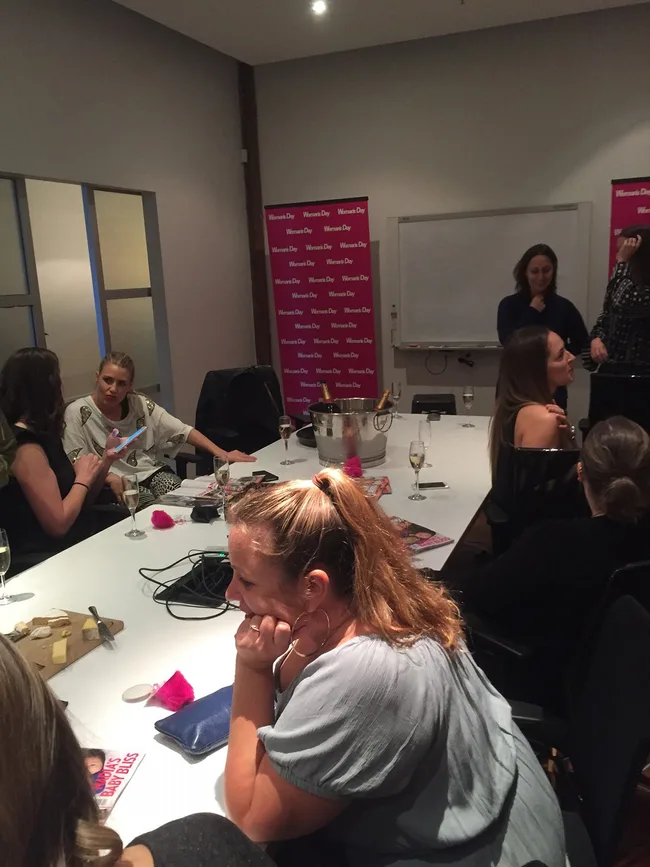 Group of women at a table with Women's Day banners in the background, discussing with drinks and magazines.