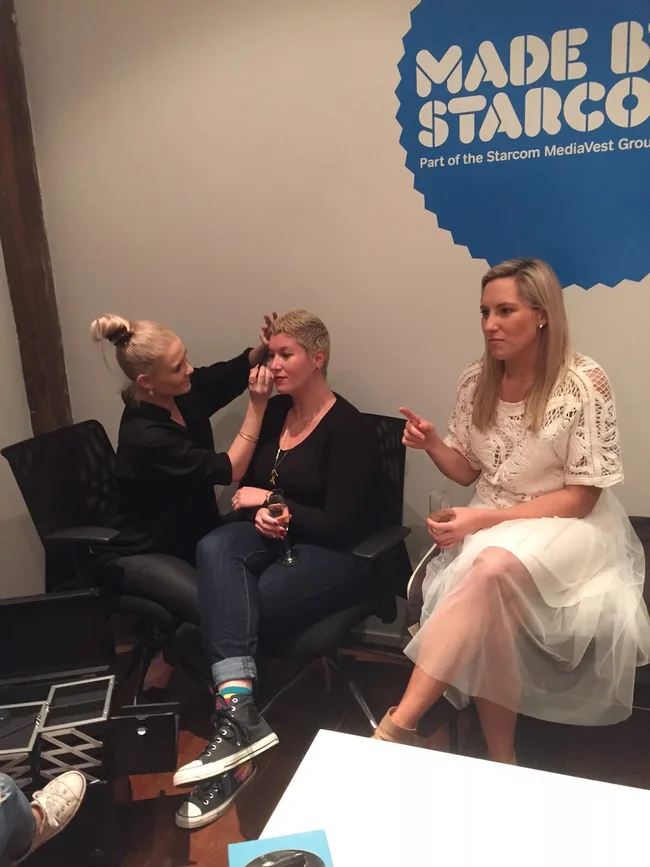 Two people seated, one doing another's makeup, third person in a white dress observing, with "Made by Starcom" logo on wall.