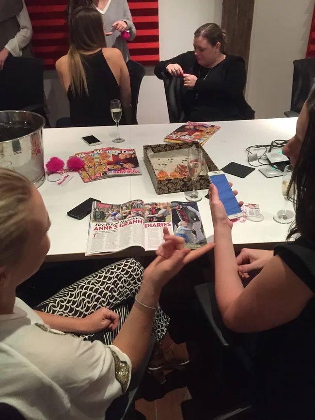 Group of people at a table with magazines, food, and drinks; one person checks a phone.