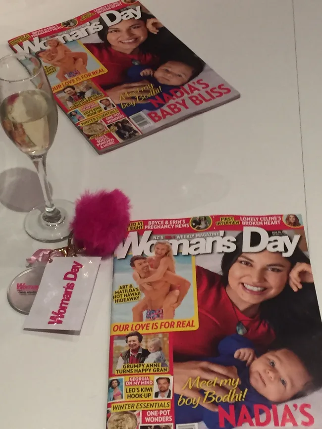 A table with Woman's Day magazines featuring a smiling woman with a baby, next to a glass of champagne and keychain.