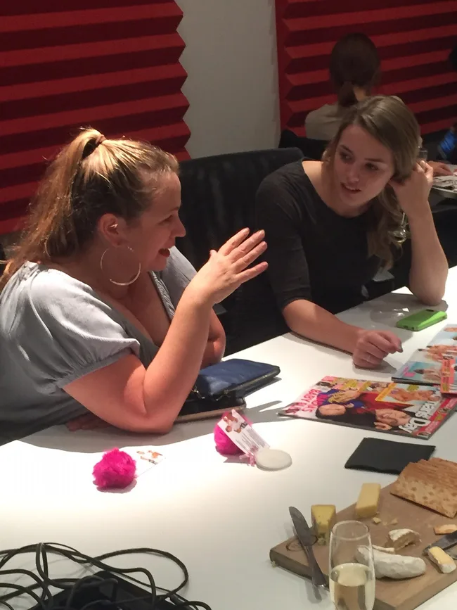 Two women talking at a table with snacks, magazines, and a glass of champagne.