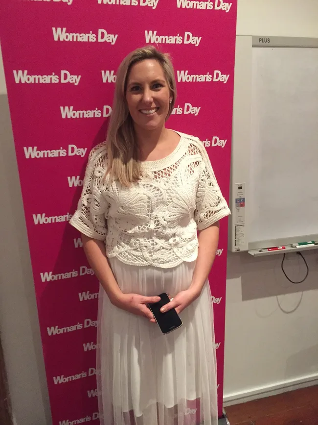 A person smiling, standing in front of a "Woman's Day" step-and-repeat backdrop, holding a phone.