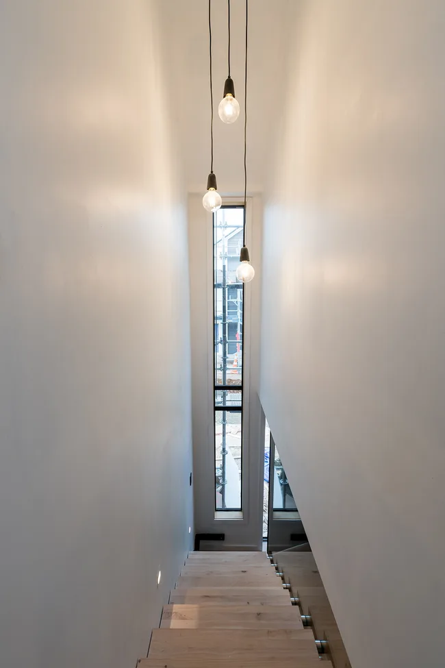 Alt text: "Minimalist staircase with wooden steps and hanging light bulbs, tall narrow window at the end."