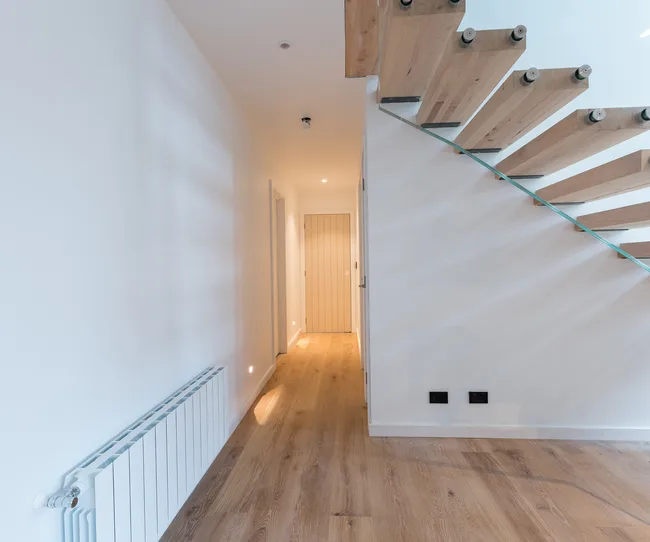 Modern interior hallway with wooden staircase, light wooden floor, white walls, and a radiator on the left.