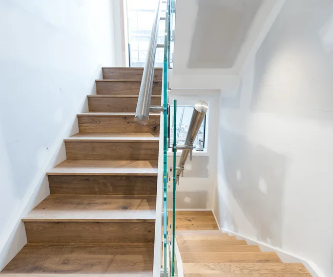 Wooden staircase with metal handrail and glass panels, in a well-lit, partially painted interior space.