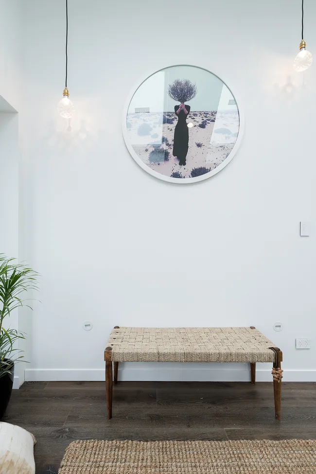Room with a bench, framed circular artwork of a figure with branches for a head, and hanging lights on the wall.