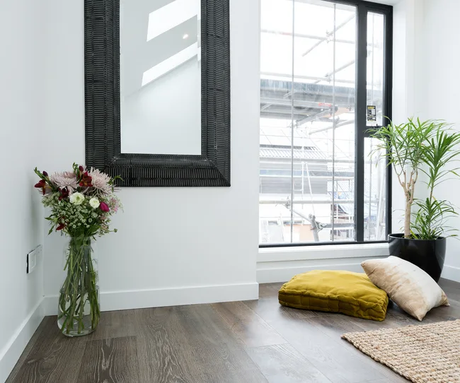 Bright room with large mirror, window, vase of flowers, cushions, and potted plant on wooden floor.