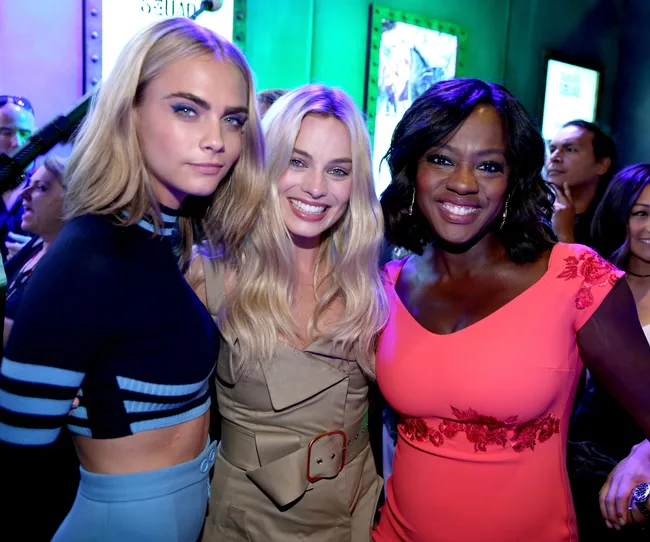 Three women posing closely together, smiling at an event with colorful lighting and a crowd in the background.