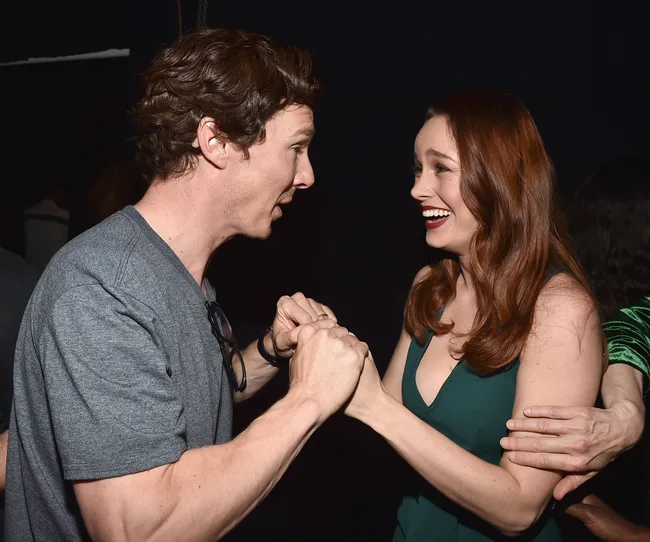 Two people warmly greet each other, clasping hands, with expressions of surprise and joy.