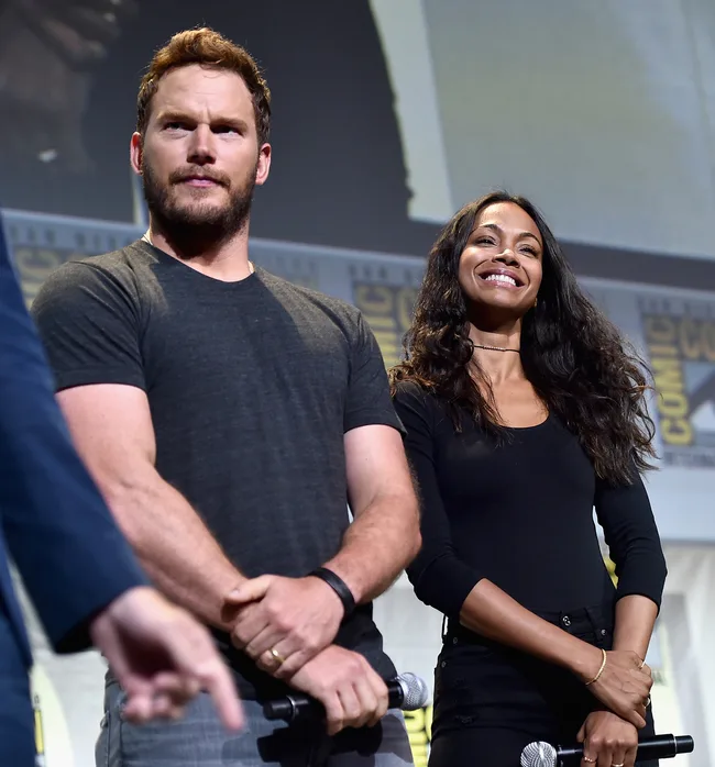 Two people standing on stage at a Comic-Con event, holding microphones, one wearing a black shirt, the other a black long-sleeve top.