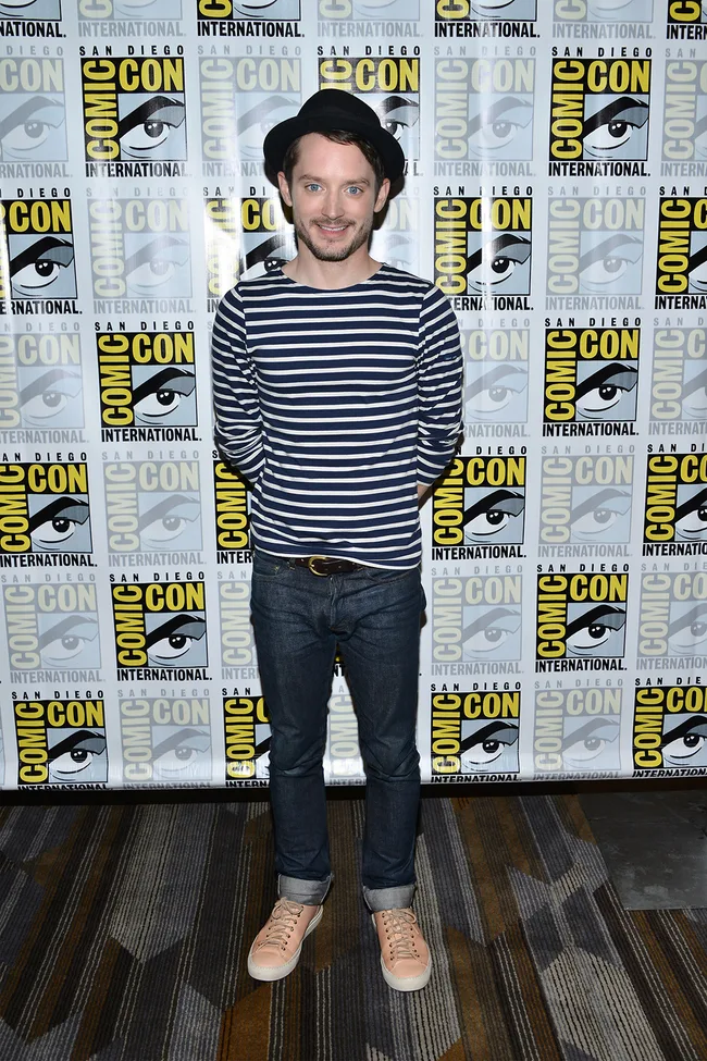 Person in a striped shirt and hat stands smiling at Comic-Con backdrop.