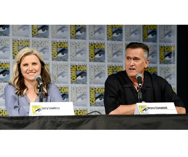 Two people, Lucy Lawless and Bruce Campbell, speak at a Comic-Con panel with microphones on a table.