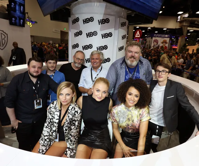 Group of people, including actors, at an HBO booth during a convention with a crowd and displays in the background.