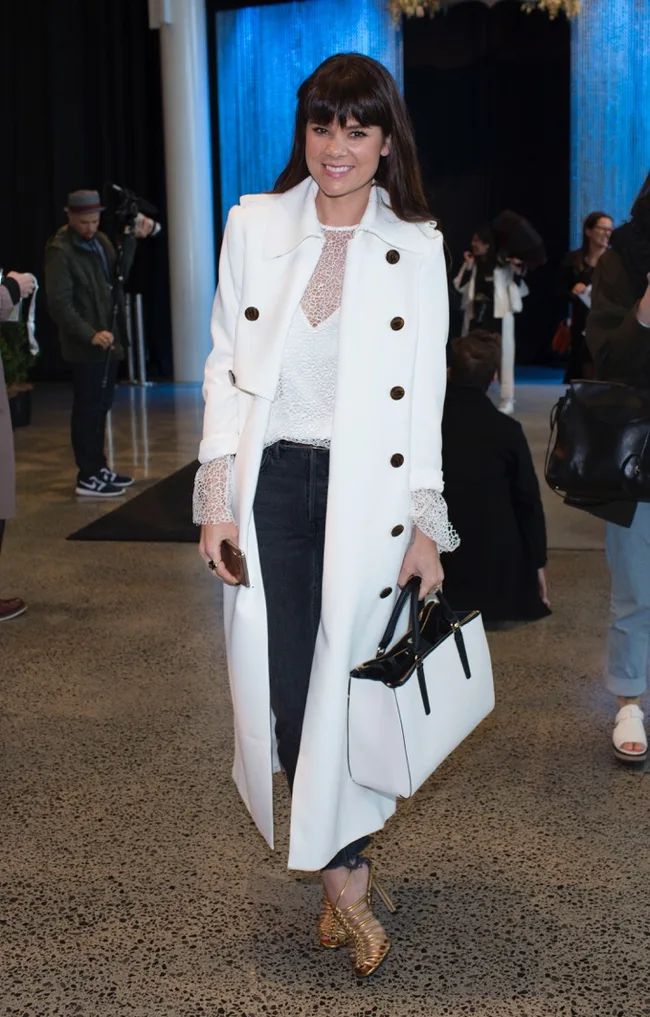 Woman in a white coat, black pants, and gold heels holding a black and white handbag, smiling at an event.