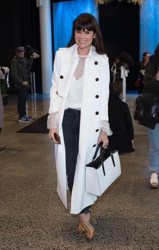 Woman in a white coat, black pants, and gold heels holding a black and white handbag, smiling at an event.