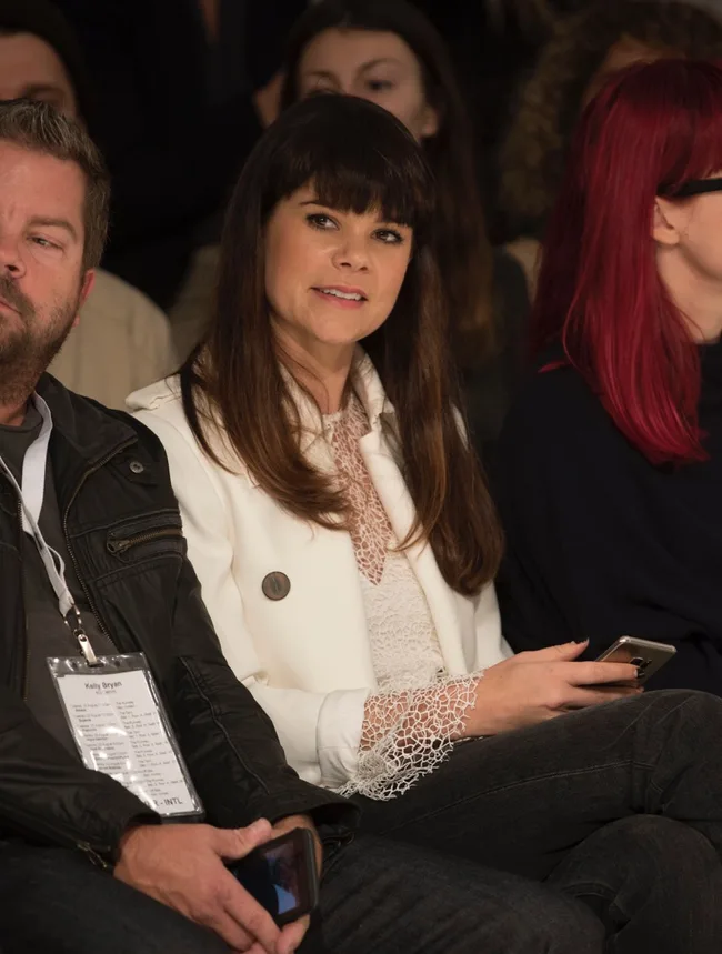 A woman in a white jacket sitting at an event, holding a phone, with others sitting nearby.