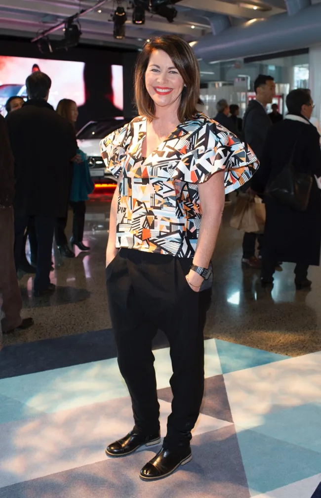 Woman in a colorful patterned top and black pants at a social event, smiling, with hands in pockets.