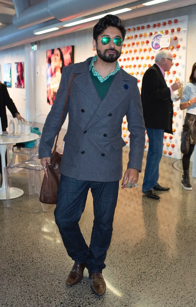 Man in sunglasses, grey blazer, and jeans stands in a modern setting with artwork on walls, holding a brown bag.