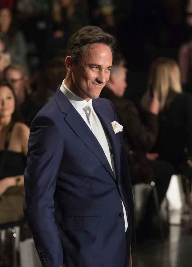 A man in a blue suit walking confidently on a runway, audience in the background.