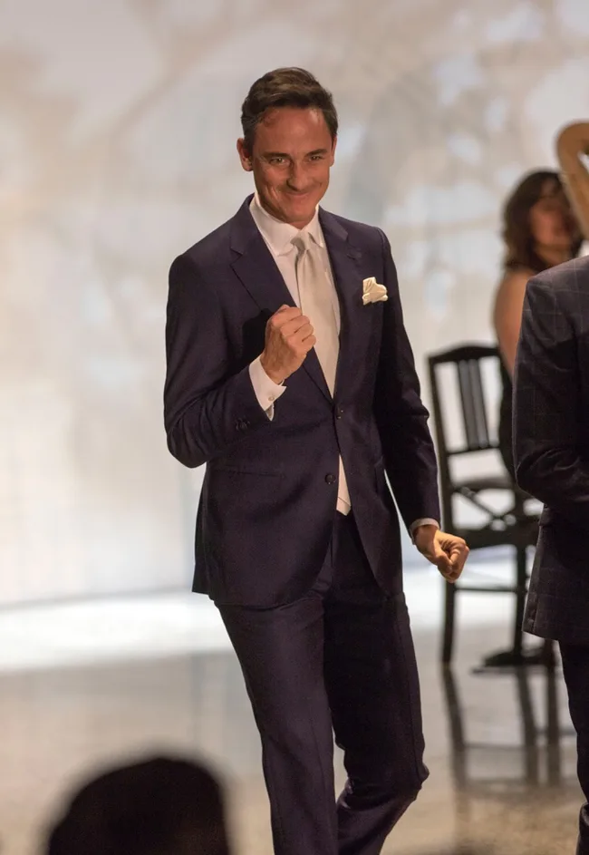 A man in a navy suit, smiling and making a fist gesture, stands in a well-lit room with chairs in the background.