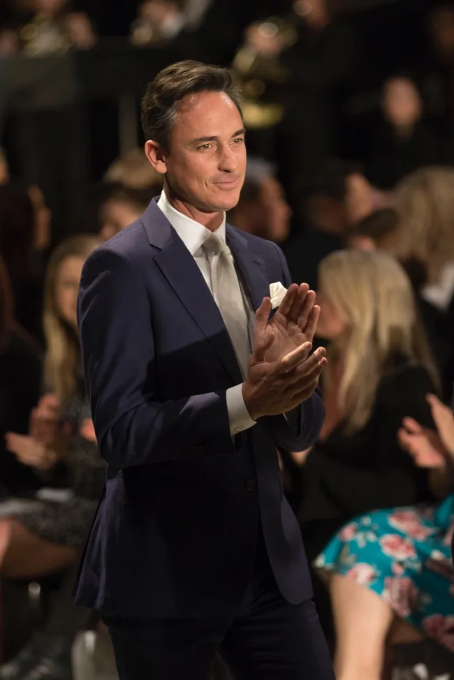 Man in a blue suit clapping in a crowded room, with others applauding in the background.